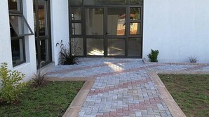 A modern building with a paved entrance area, large glass doors, and a small garden with plants. - Tsumeb Guesthouse Kamho (Tsumeb)