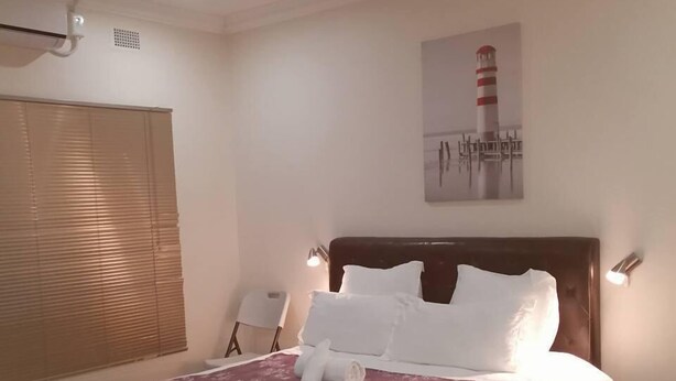 A neatly made bed with white linens and a red throw, a nightstand with a lamp, a chair, and a painting of a lighthouse on the wall. - Tsumeb Guesthouse Kamho (Tsumeb)