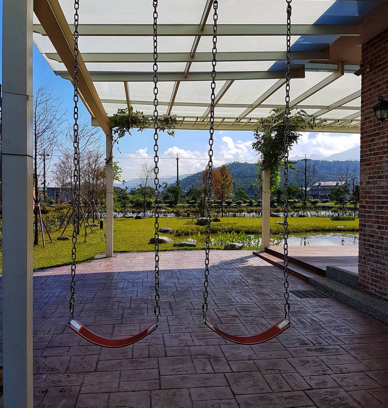 A covered outdoor area with a swing set, brick flooring, and a view of a landscaped garden with a pond and distant mountains.