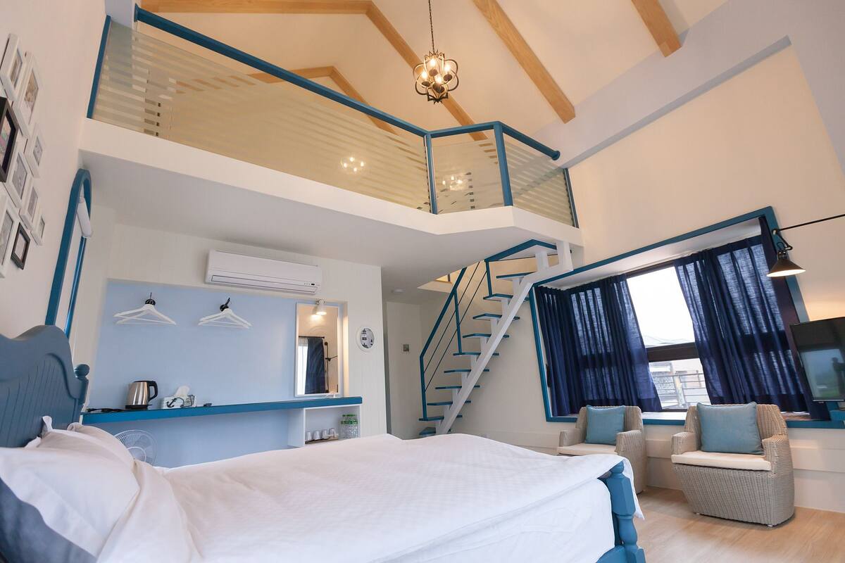 A modern bedroom with a large bed, a staircase to an upper level, a seating area with chairs, and a television.