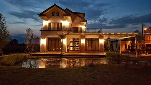 A two-story house with a large front porch, a covered outdoor seating area, and a pond in the foreground. - More Manor (Dongshan)