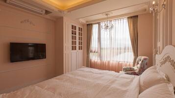 A bedroom with a large bed, a television mounted on the wall, a window with curtains, and a chandelier.