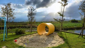 A playground with a yellow slide and a blue swing set in a grassy area with trees and a river in the background. - More Manor (Dongshan)