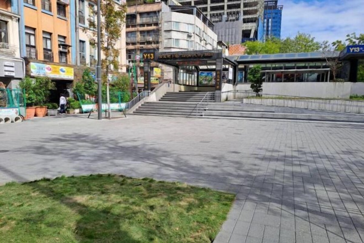 A public square with a staircase leading to a covered area, surrounded by buildings and greenery.