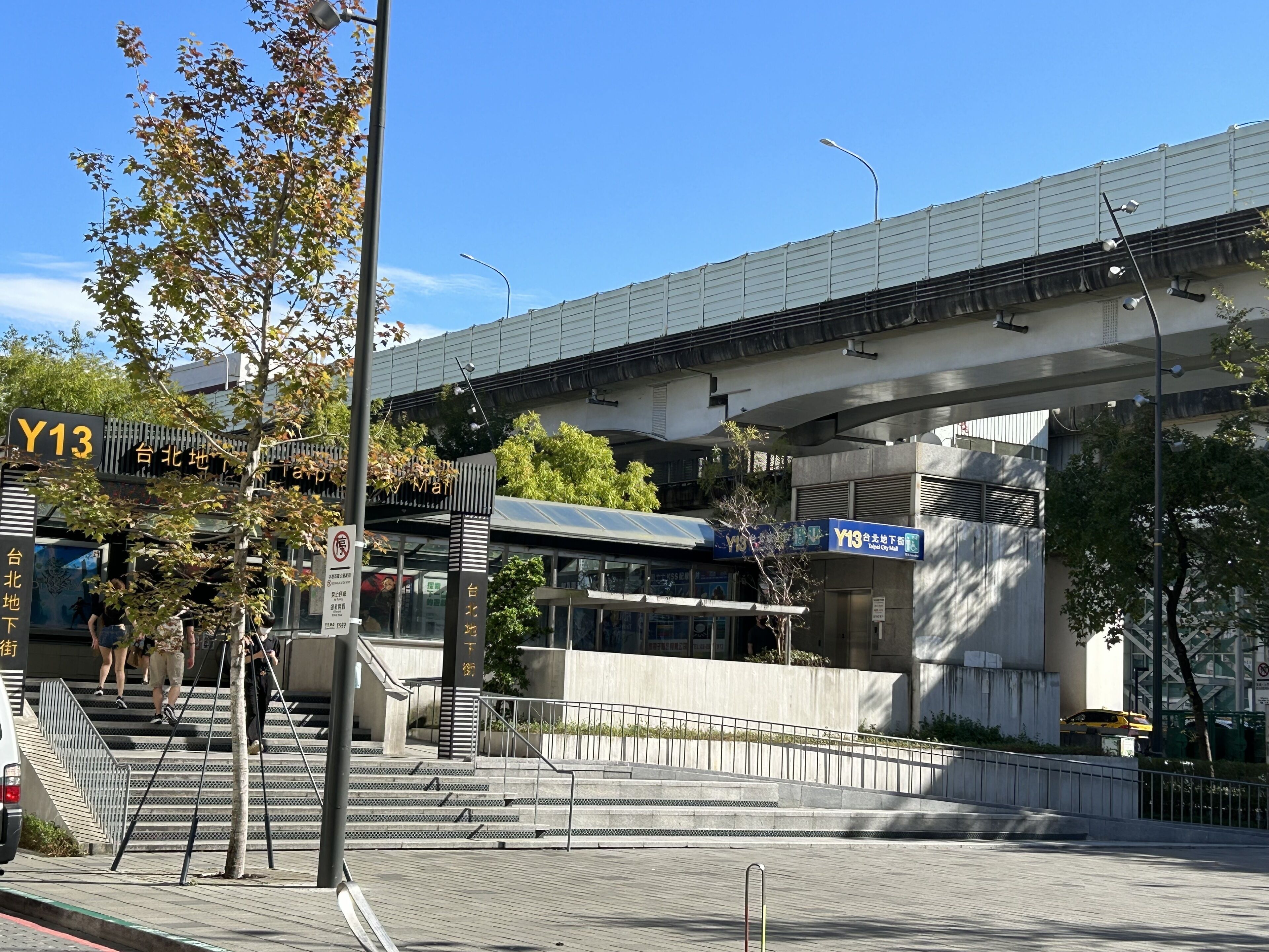 A modern urban area with a pedestrian walkway, a covered bridge, and signage indicating a location.