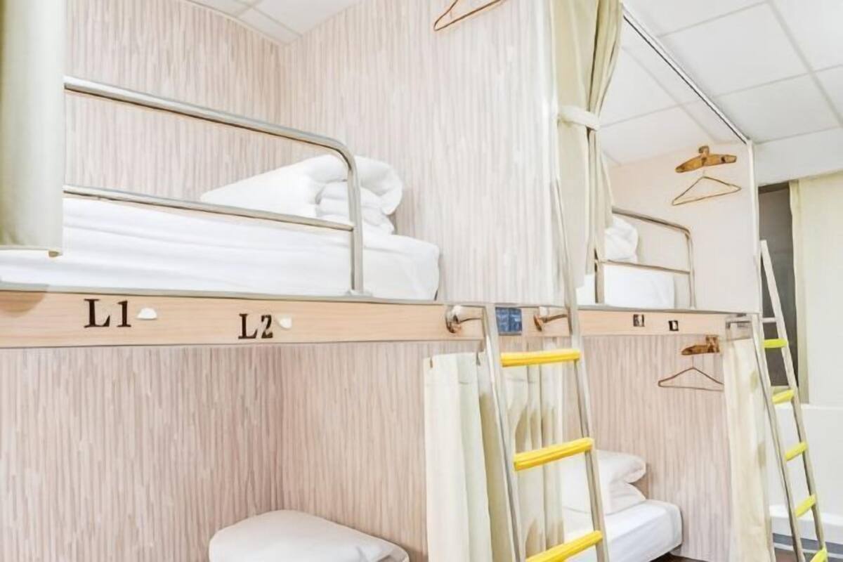 Ein Mehrbettzimmer mit zwei beschrifteten Betten, L1 und L2, einem Nachttisch, einem Hängesessel und einer Leiter.