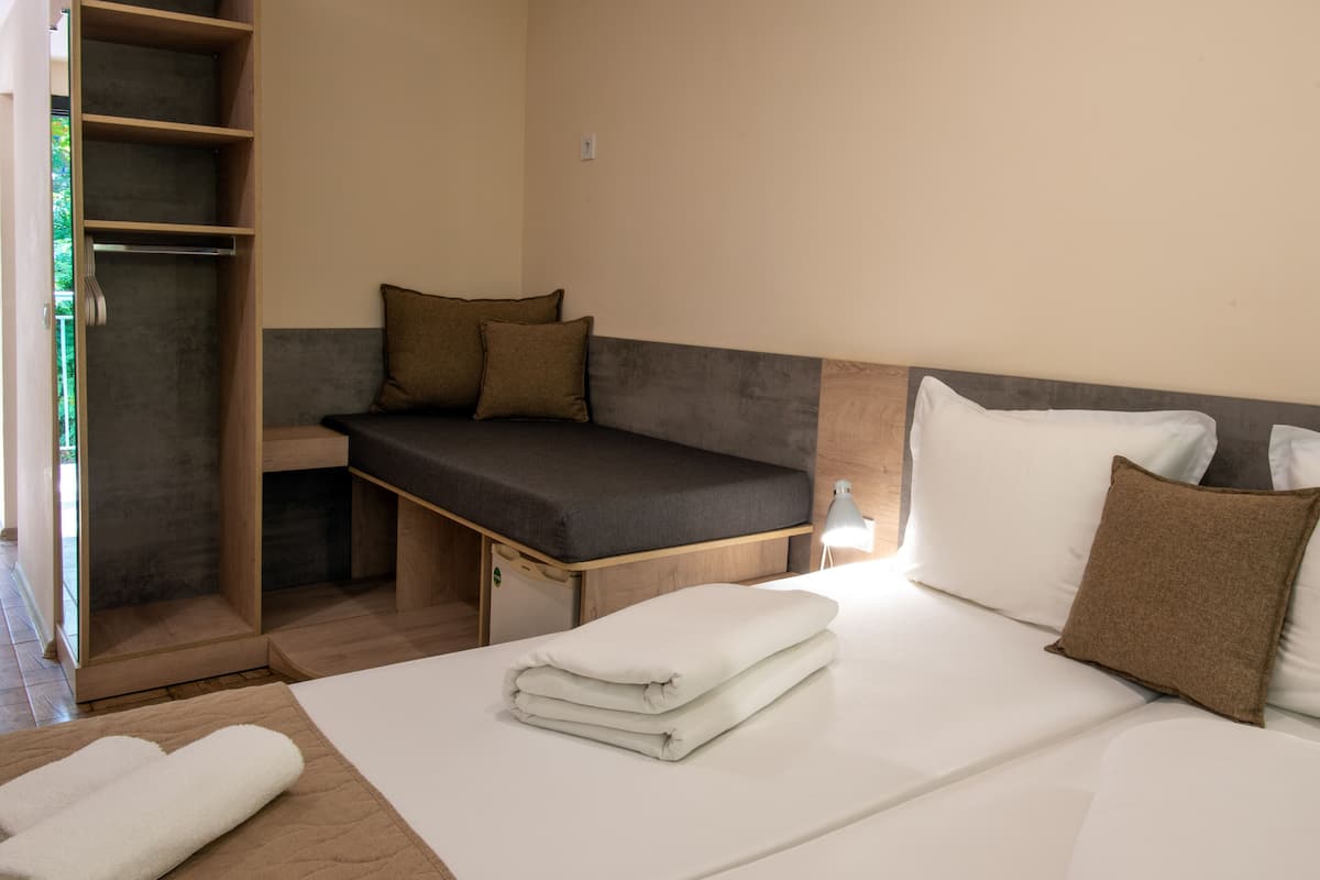 A compact hotel room with a bed, a small seating area, a wardrobe, and a bedside table with a lamp.
