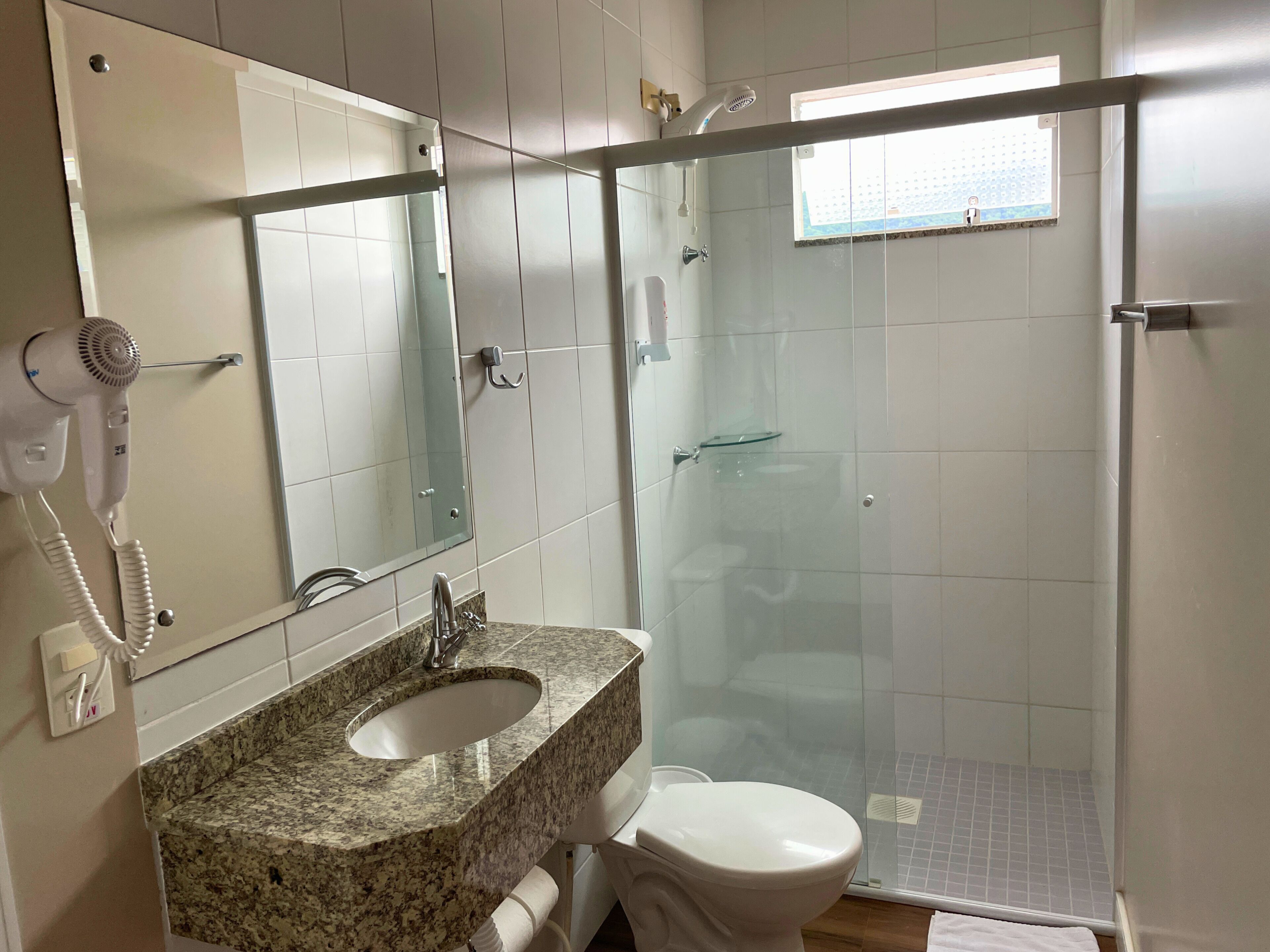 A bathroom with a granite countertop sink, a white toilet, a glass shower enclosure, and a hairdryer mounted on the wall.