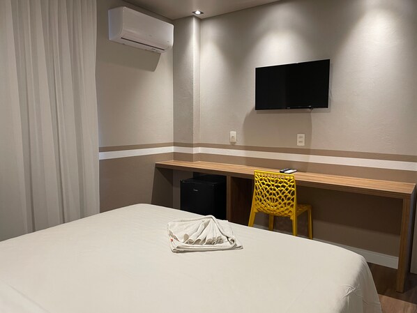 Una habitación de hotel con cama, escritorio, silla, aire acondicionado y televisor montado en la pared.