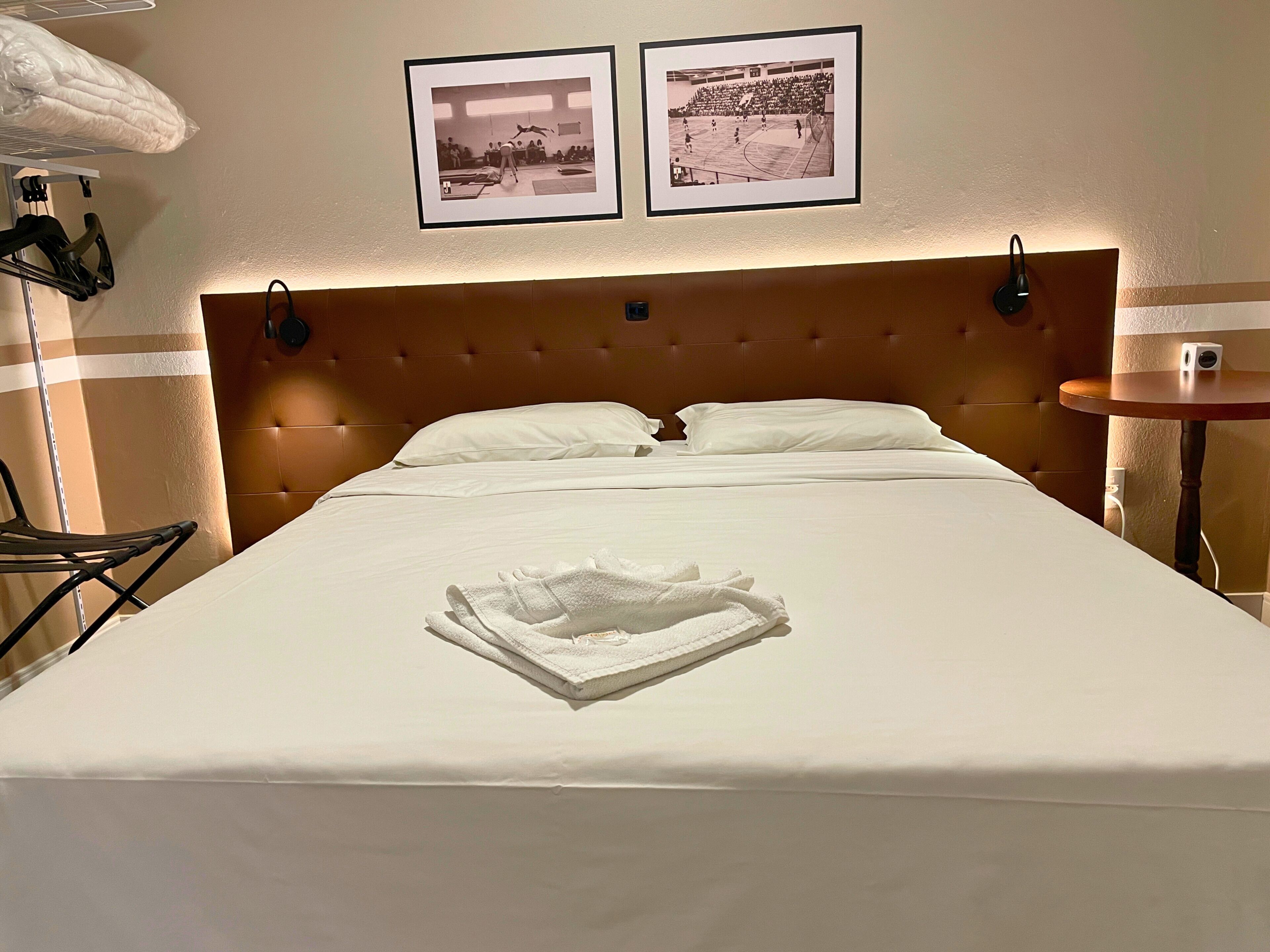 Quarto de hotel com uma cama grande, duas fotografias emolduradas na parede, uma mesa redonda pequena e uma cadeira.