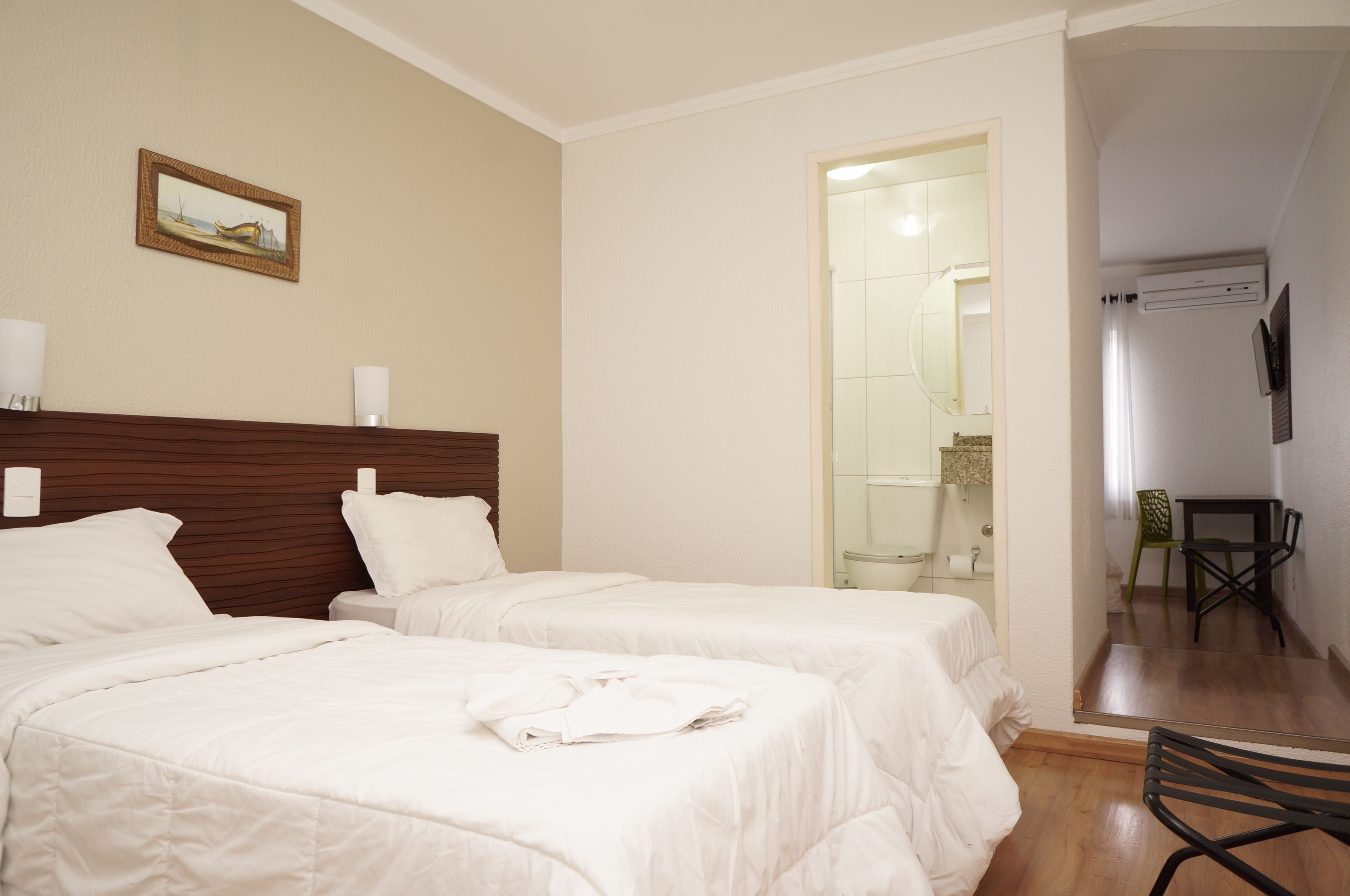 Habitación de hotel con dos camas, cabecero de madera, un cuadro en la pared, baño con puerta de cristal y una pequeña mesa con sillas.