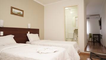 A hotel room with two beds, a wooden headboard, a painting on the wall, a bathroom with a glass door, and a small table with chairs.