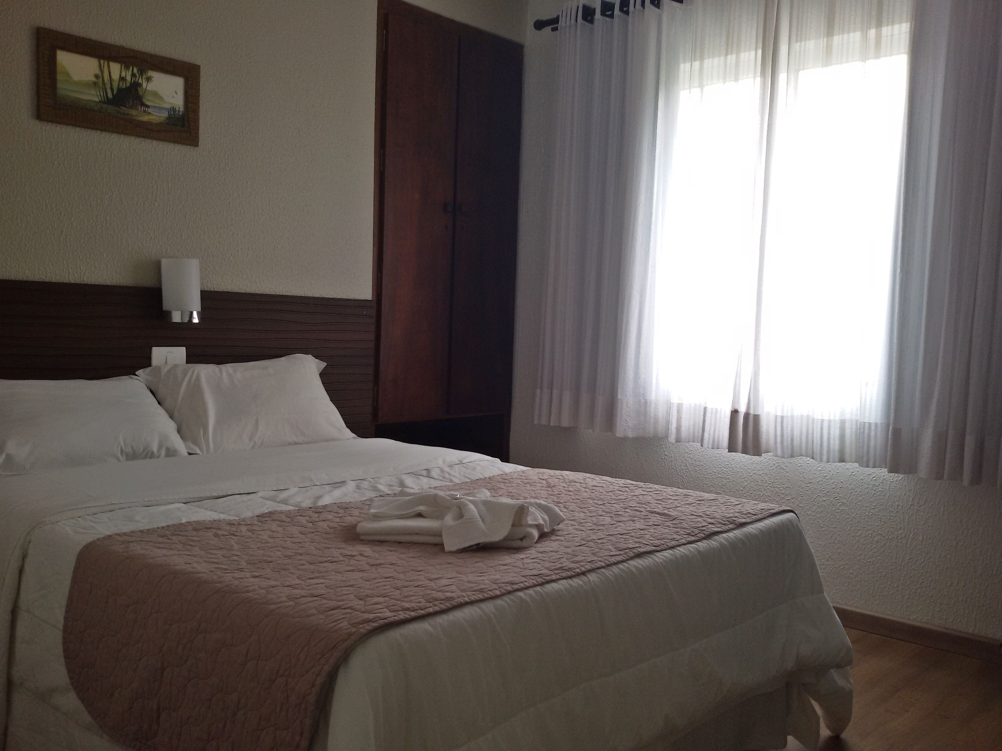 A neatly made bed with a pink quilt, white pillows, and a folded towel on the bed. There is a wooden headboard, a wall-mounted light, and a painting above the bed.
