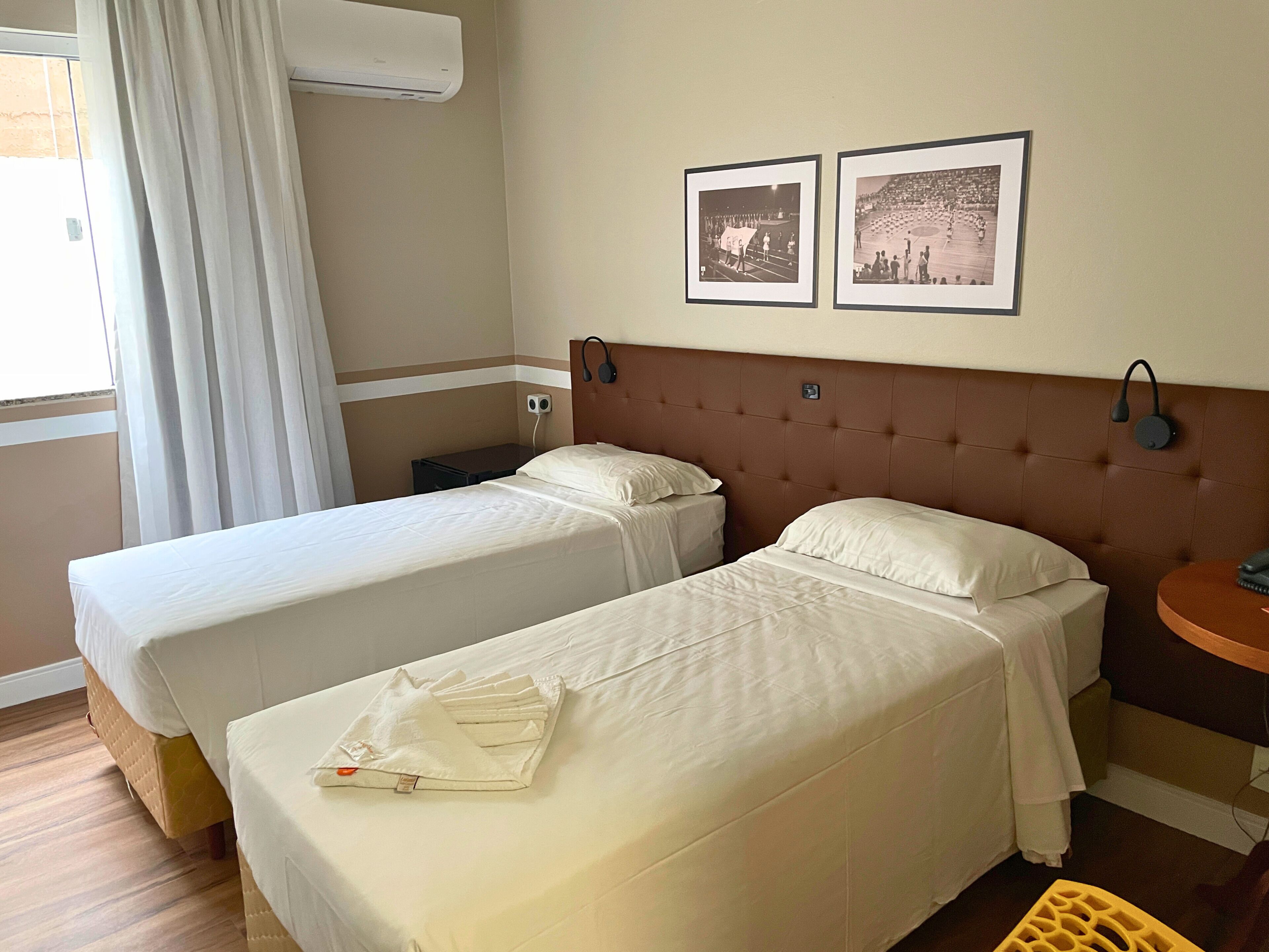 A hotel room with two single beds, a small bedside table, a wall-mounted air conditioner, and two framed pictures on the wall.