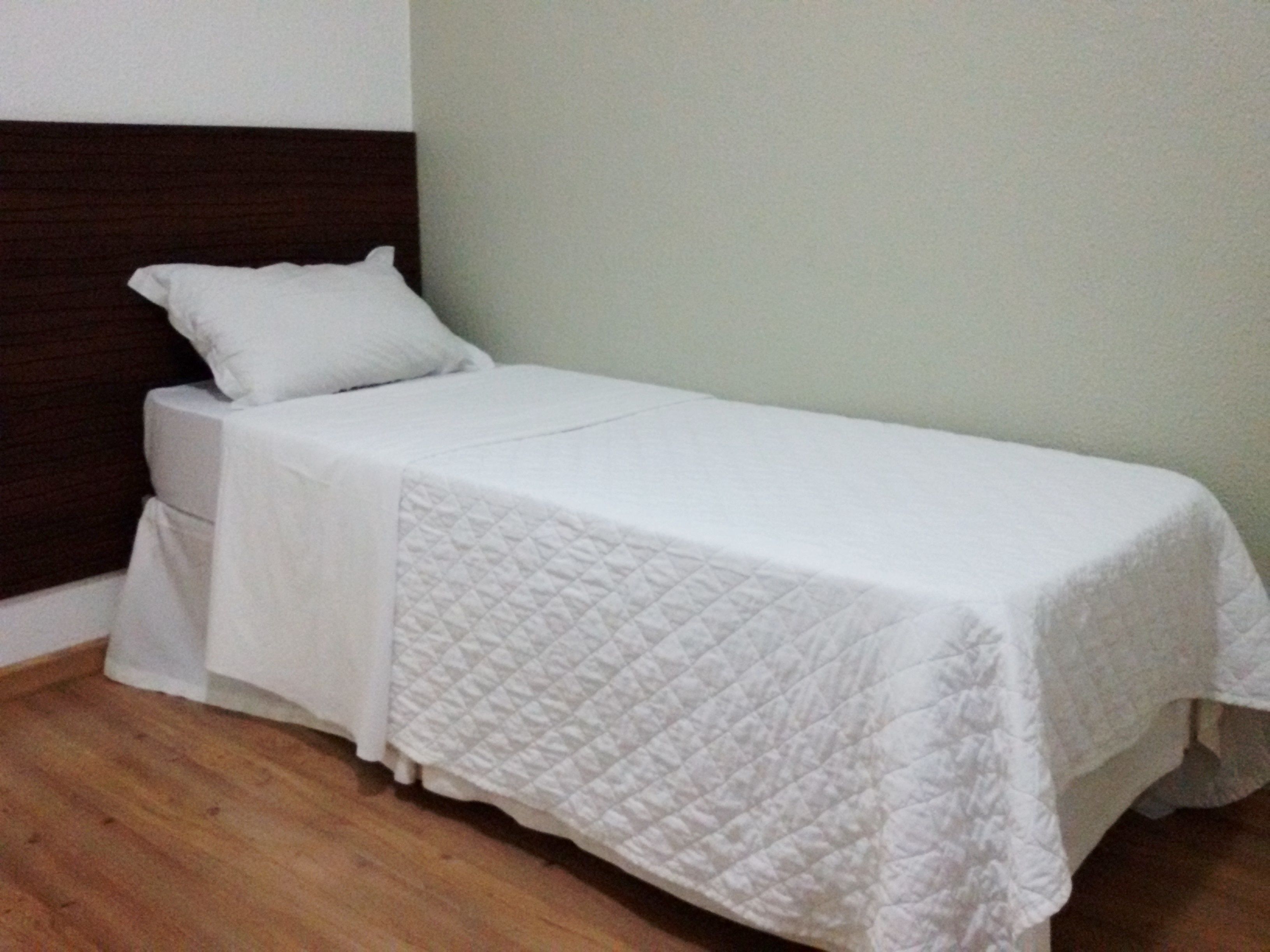 Una cama individual con una colcha blanca acolchada y almohada a juego, ubicada frente a una pared de color claro y con cabecera oscura.