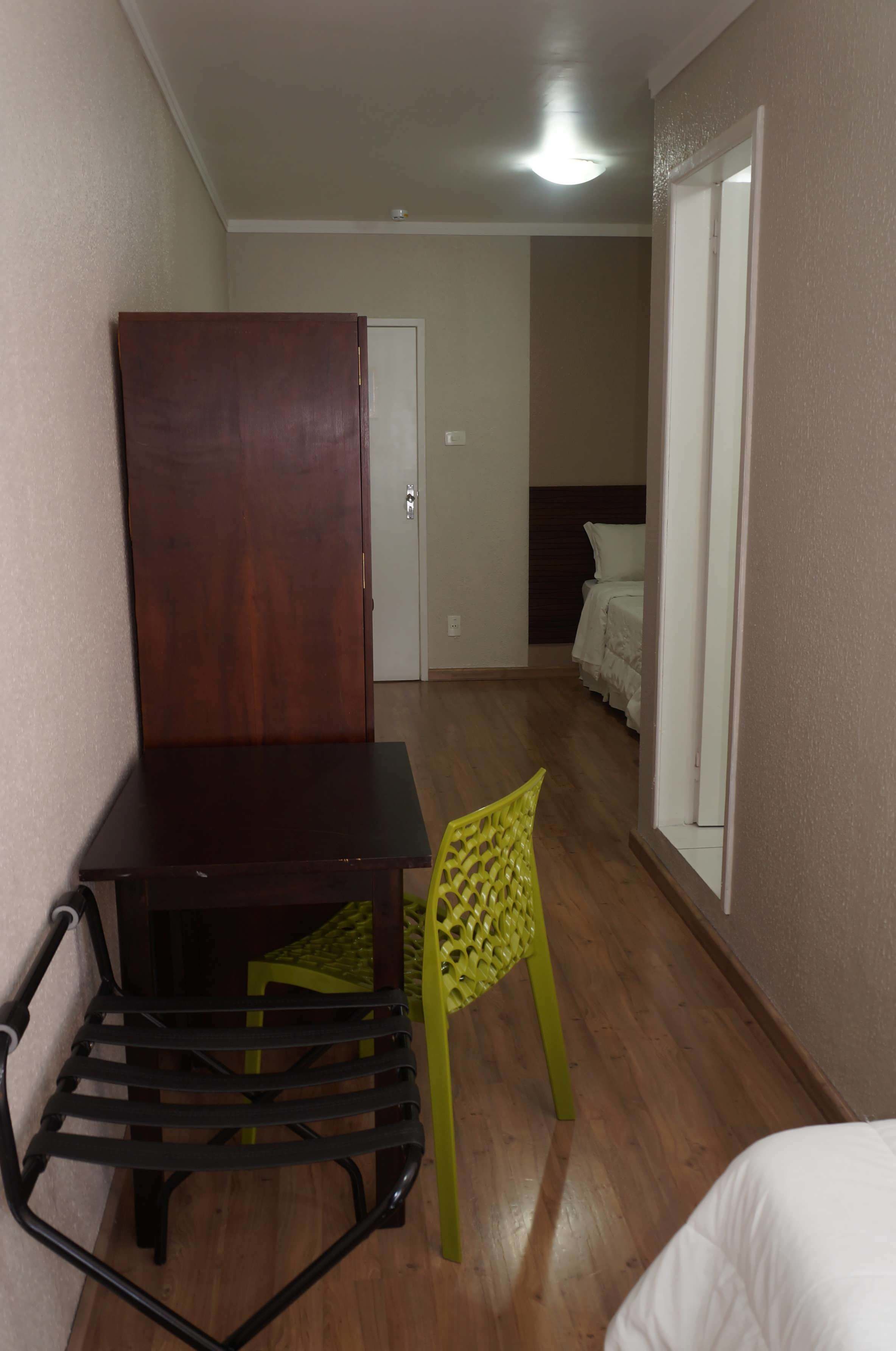 A room with a wooden desk, a green chair, a bed with white bedding, and a door leading to another room.