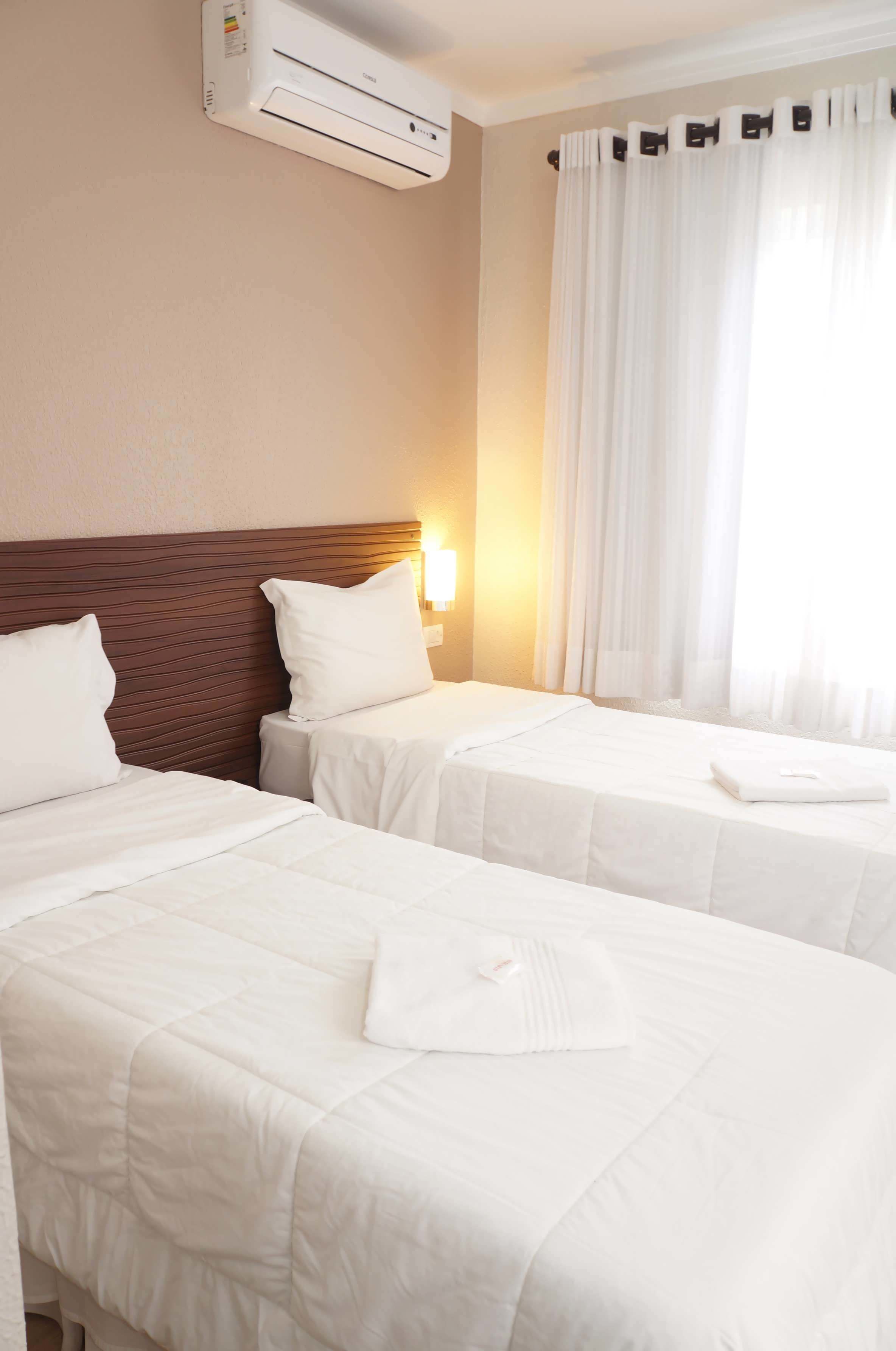 Habitación de hotel con dos camas, aire acondicionado de pared, luz nocturna y una ventana con cortinas blancas.