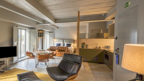 Un salon moderne avec un canapé, une table basse et un téléviseur. Il y a également un coin repas avec des chaises et une table. La pièce est dotée de grandes fenêtres et offre une vue sur l’extérieur.