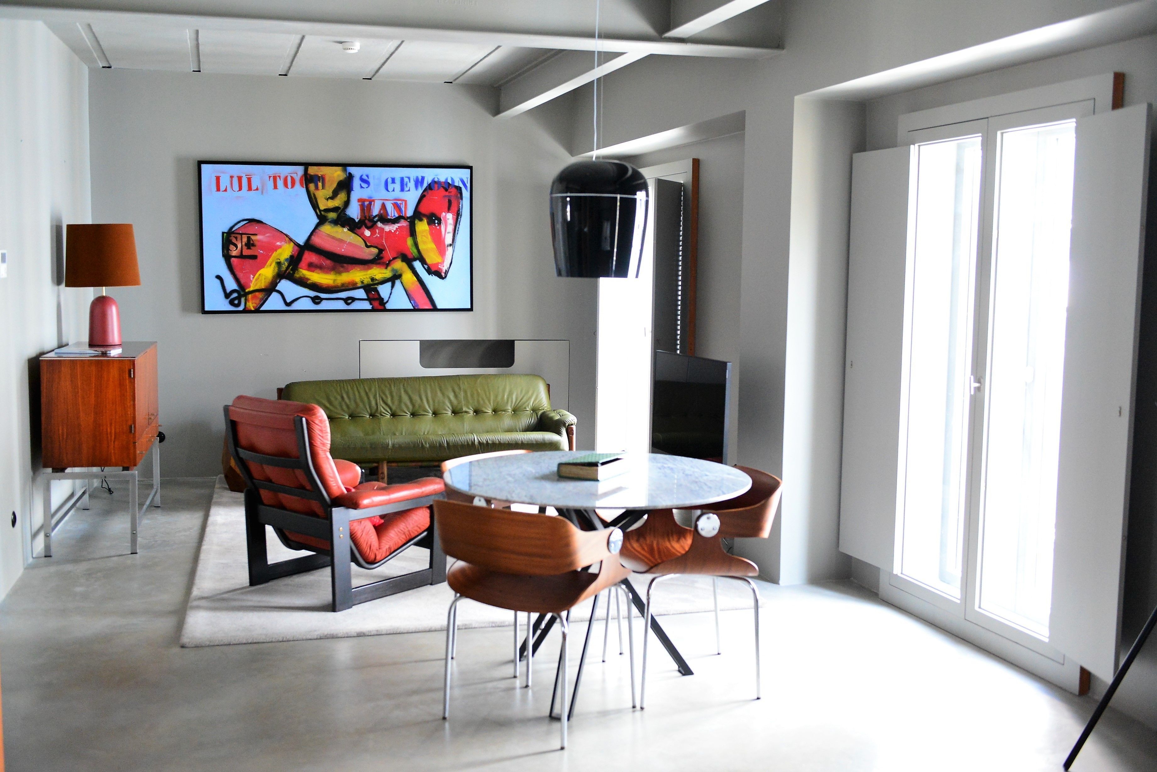 A modern living room with a green sofa, red and brown chairs, a round table, and a large abstract painting on the wall.