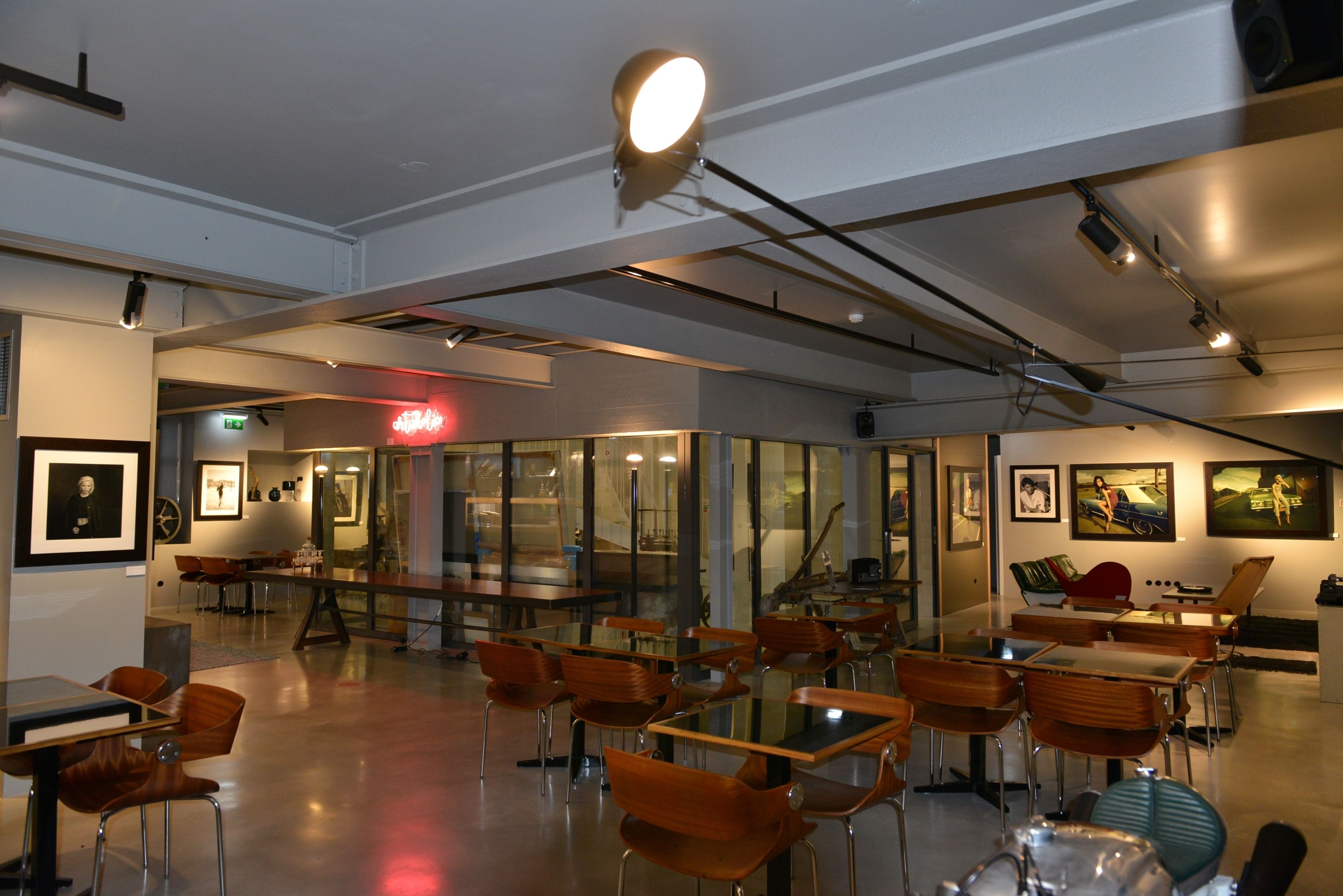 A modern dining area with glass tables and wooden chairs, framed artwork on the walls, and a bright ceiling light.