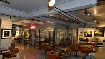 A modern dining area with glass tables and wooden chairs, framed artwork on the walls, and a bright ceiling light.