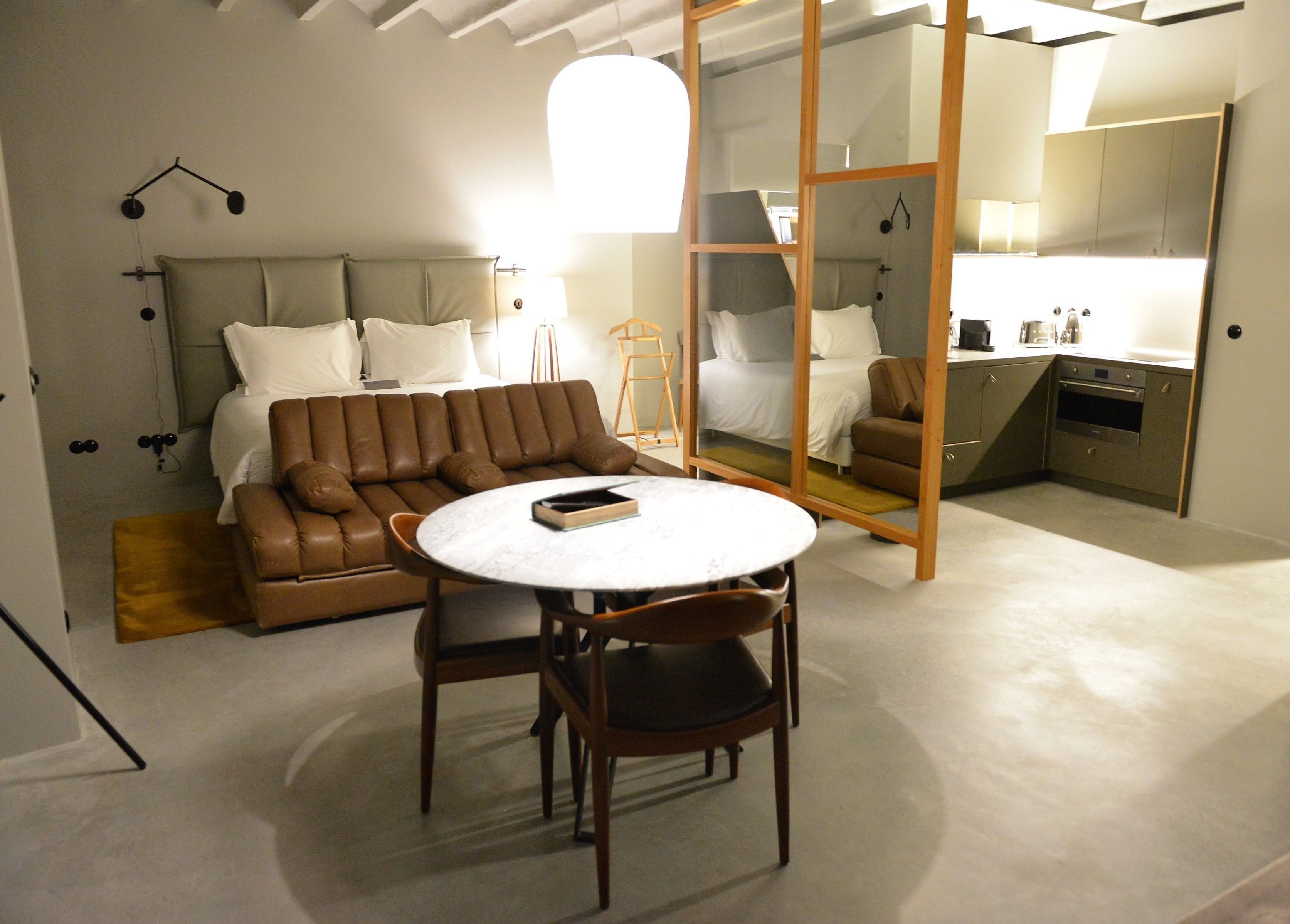 A modern bedroom with a large bed, a brown leather sofa, a round table, and a small kitchen area.