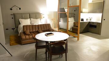 A modern bedroom with a large bed, a brown leather sofa, a round table, and a small kitchen area.
