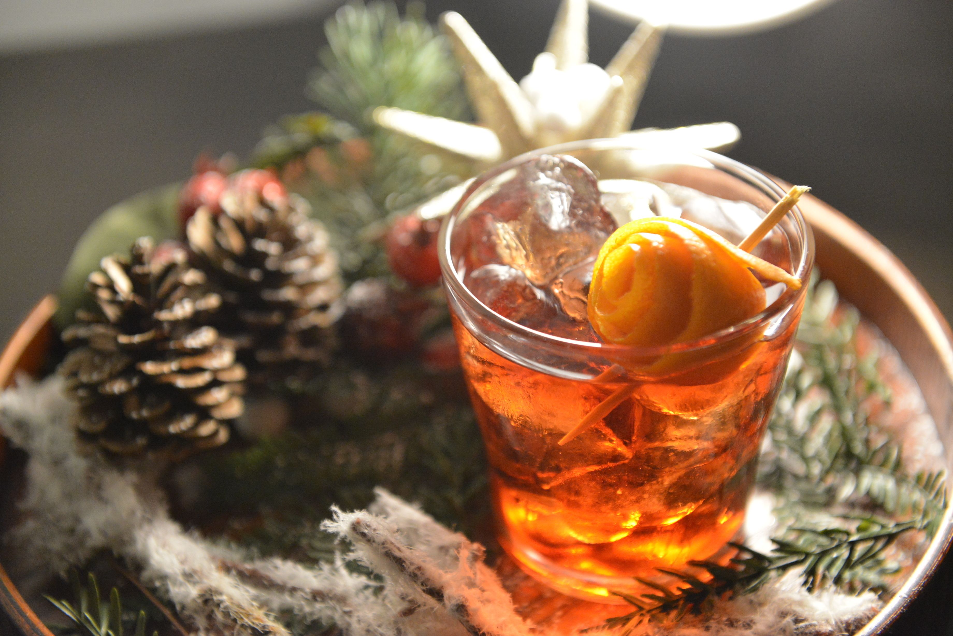 A glass of amber liquid with a garnish, surrounded by pine cones and greenery, suggesting a festive or holiday setting.