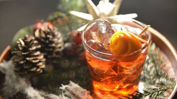 A glass of amber liquid with a garnish, surrounded by pine cones and greenery, suggesting a festive or holiday setting.
