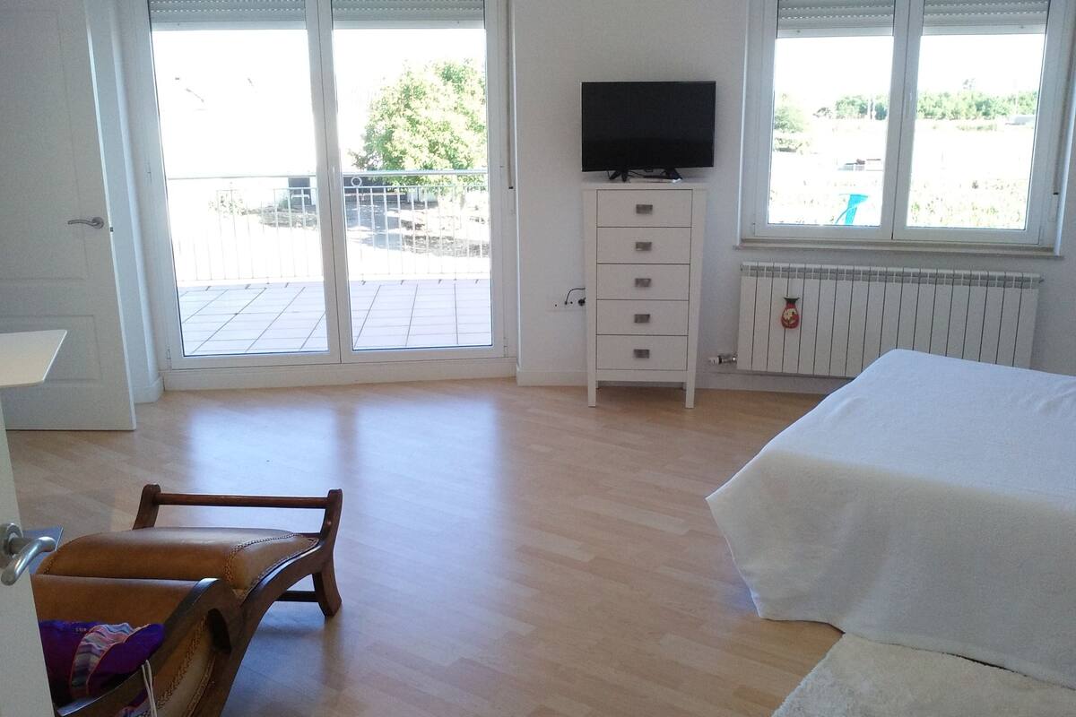 A bedroom with a bed, a television on a stand, a wooden chair, a white dresser, and a view of the outdoors through large windows.