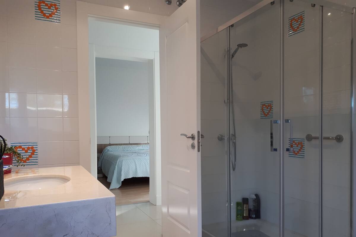 A bathroom with a marble sink, a glass shower enclosure, and a bed in the adjacent room.