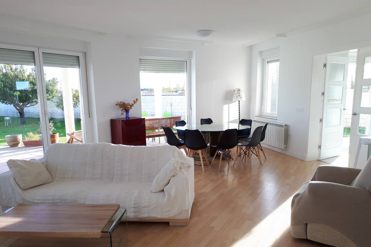 A spacious living area with a sofa, a coffee table, and a dining table with chairs. There is a large window providing natural light and a view of the outdoors.