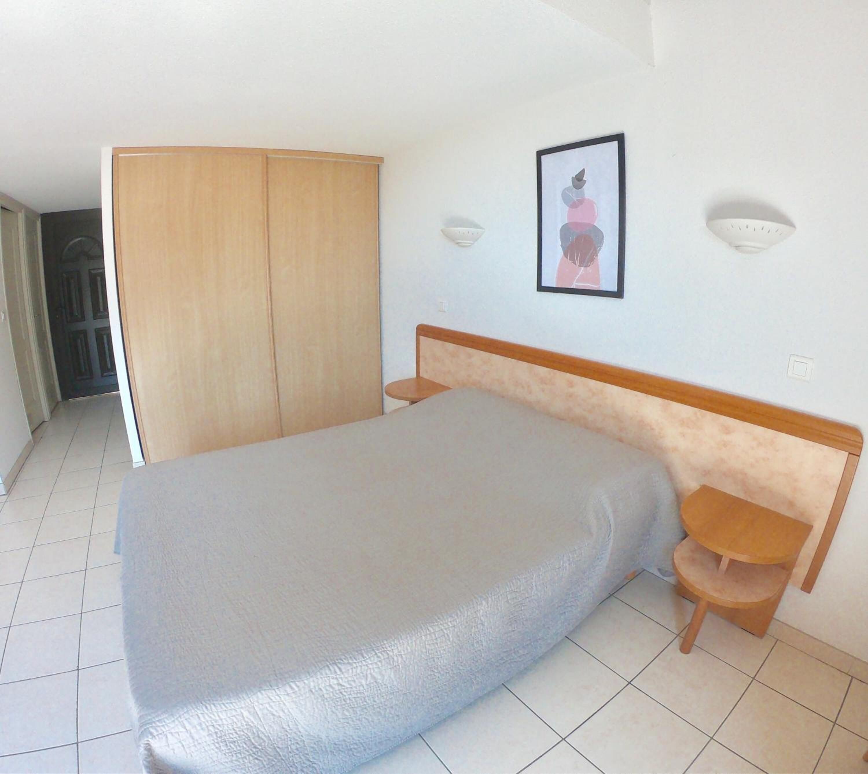 Ein Schlafzimmer mit Bett, Kleiderschrank, kleinem Tisch und Wandleuchte.