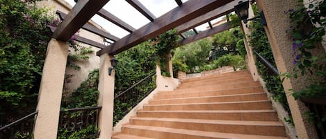A staircase with a wooden pergola overhead, surrounded by greenery and a stone wall.