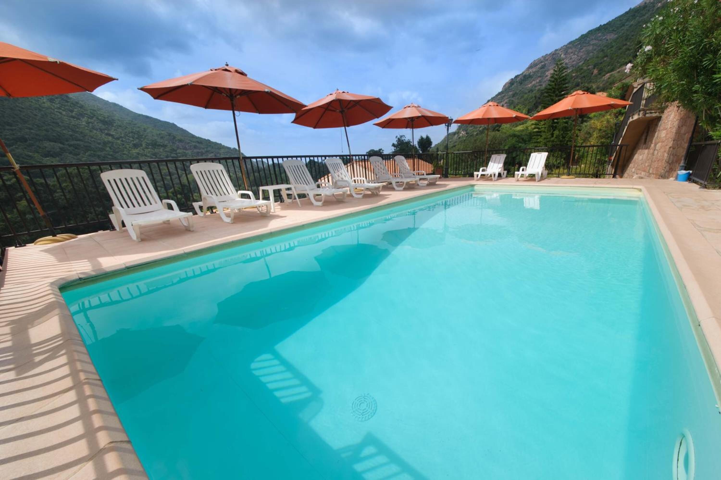 Una piscina con sillones blancos y sombrillas rojas, rodeada por un muro de piedra y vegetación.
