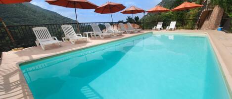 Una piscina con sillones blancos y sombrillas rojas, rodeada por un muro de piedra y vegetación.
