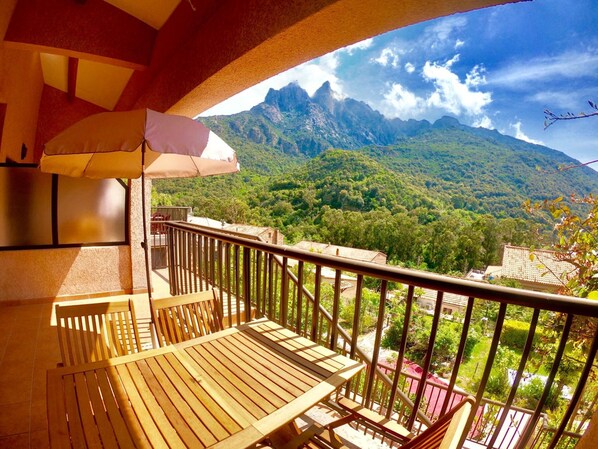 Un balcón con mesa y sillas de madera, un paraguas color beige y vistas a un paisaje montañoso.
