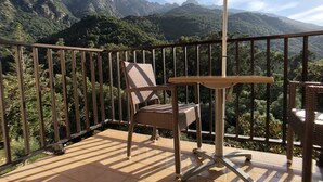 Een balkon met een rieten stoel en een tafeltje, met uitzicht op een bergachtig landschap.