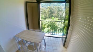 A small balcony with a table and chairs, a view of a forested area, and a sliding door.