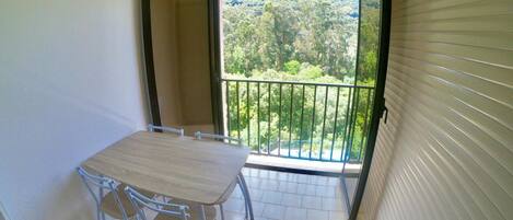 A small balcony with a table and chairs, a view of a forested area, and a sliding door.