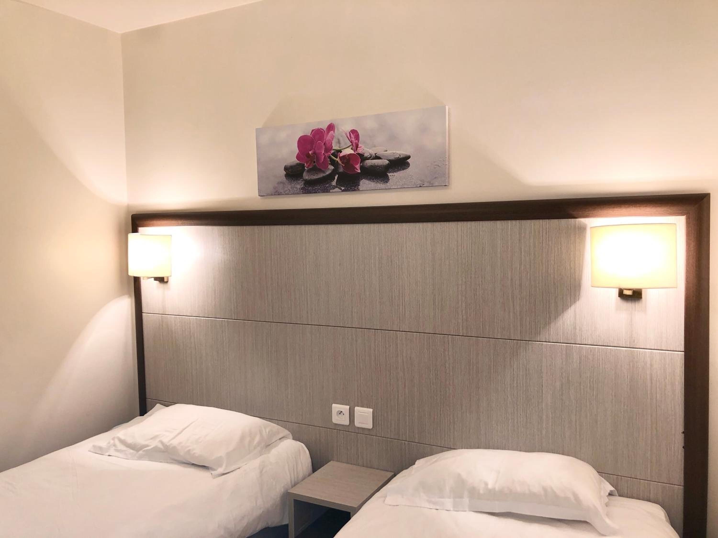 A hotel room with two beds, a nightstand, and a wall-mounted picture of pink flowers.