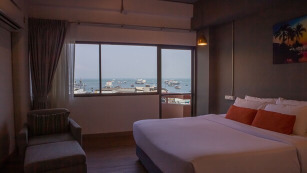 Camera d'albergo con un letto grande, una sedia, vista sul mare e sulle barche e un quadro appeso al muro.