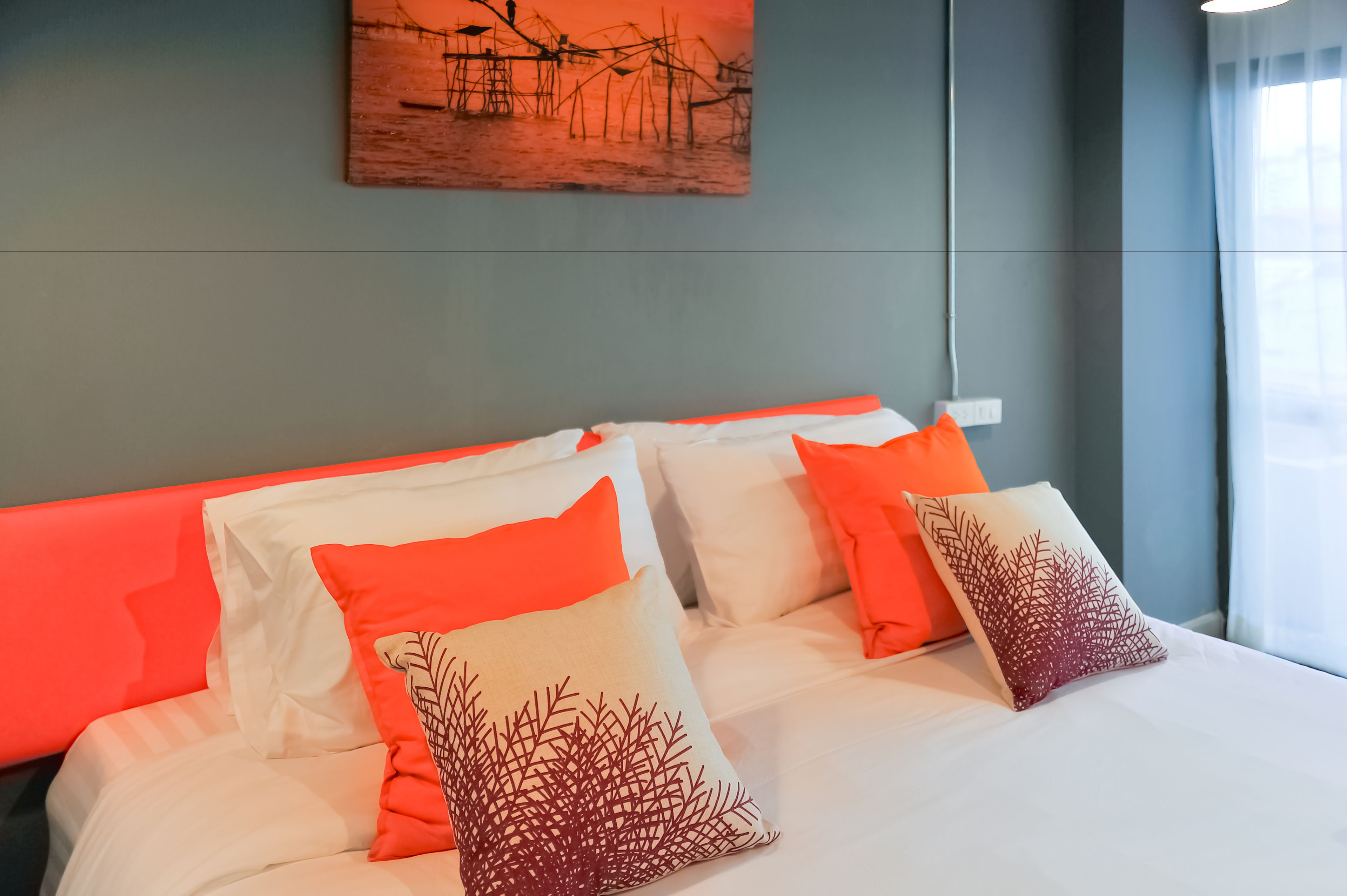 A neatly made bed with white and orange pillows, a grey headboard, and a painting on the wall.