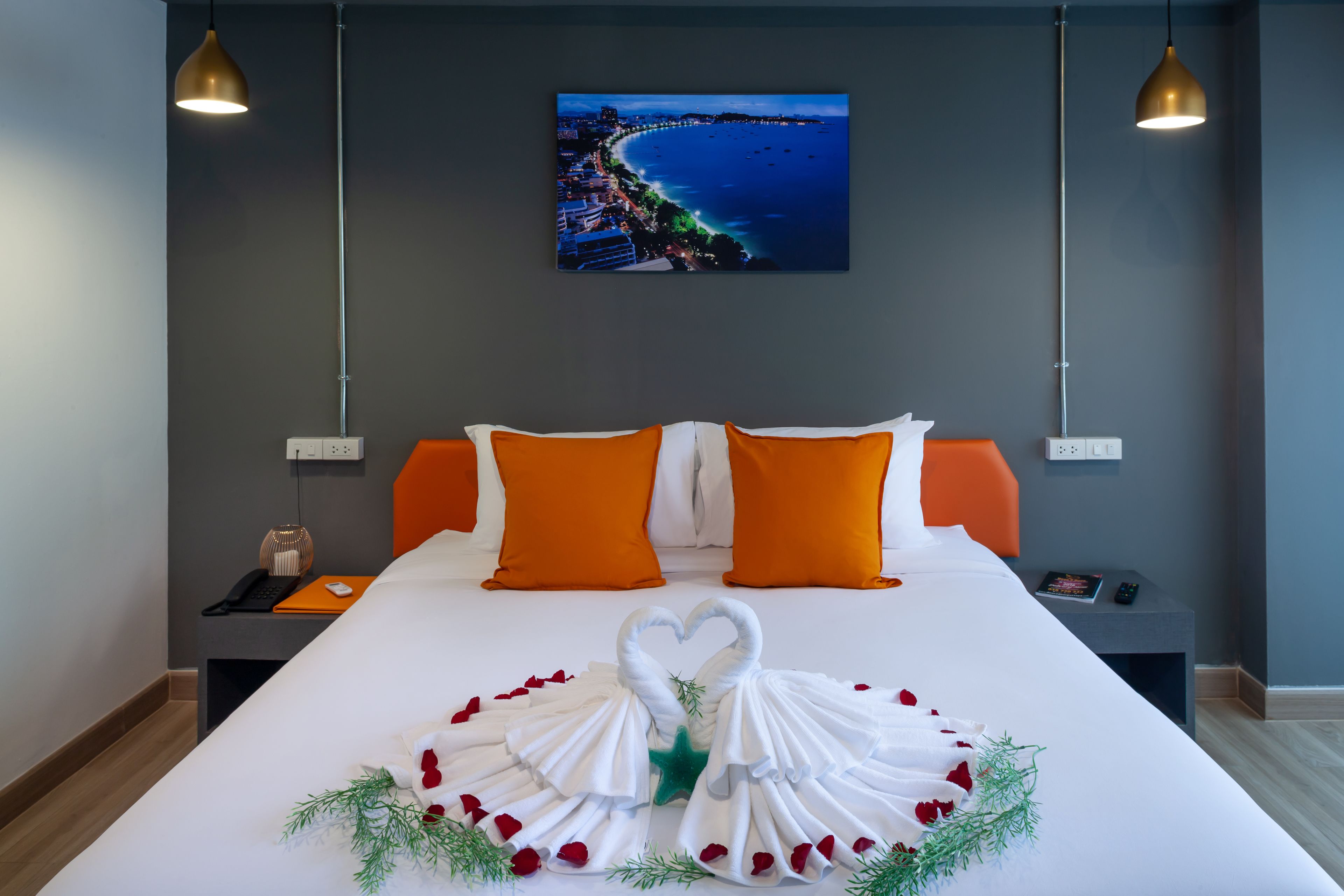 A hotel room with a large bed, orange pillows, a heart-shaped towel arrangement, and a cityscape painting on the wall.