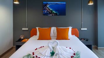 A hotel room with a large bed, orange pillows, a heart-shaped towel arrangement, and a cityscape painting on the wall.