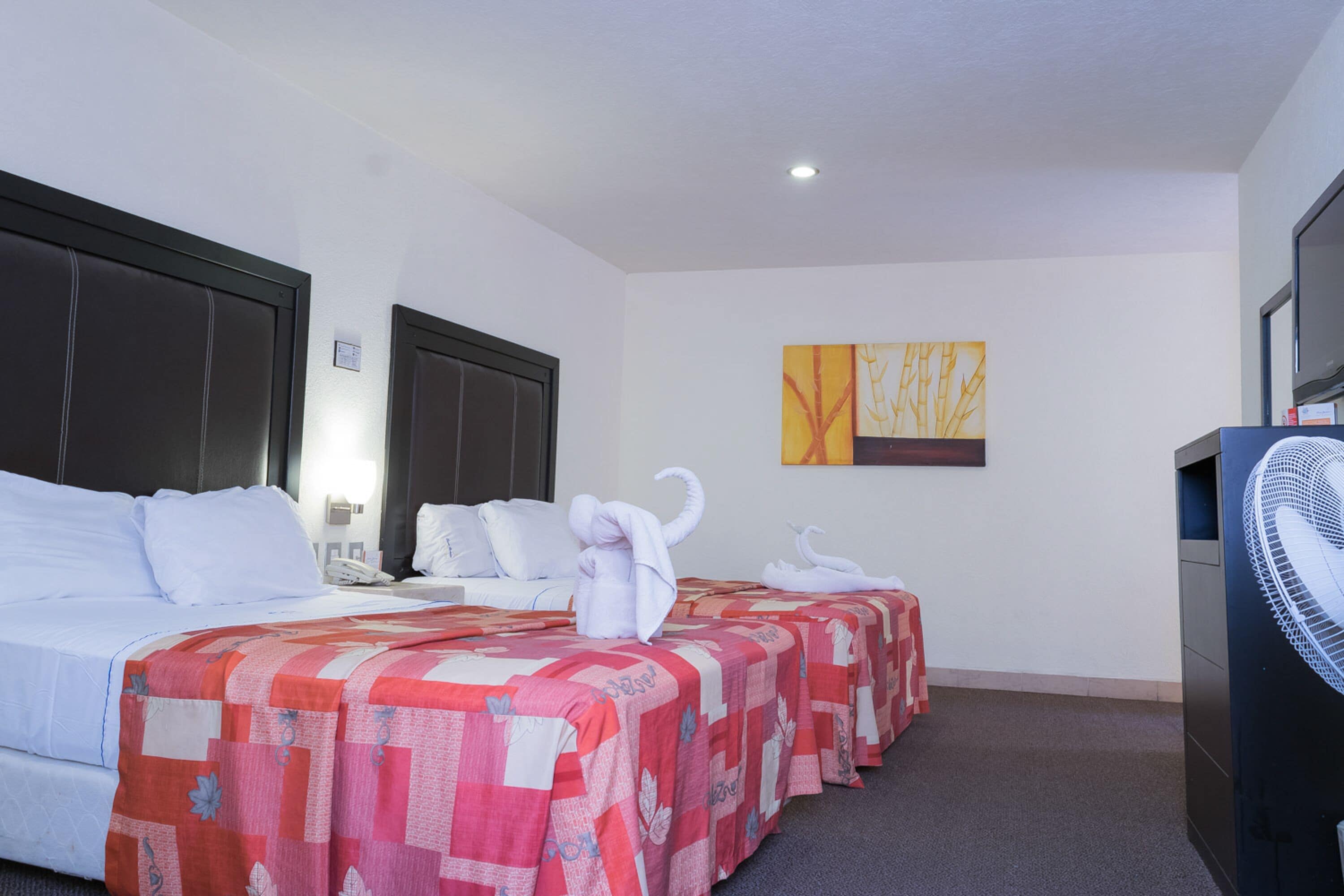 A hotel room with two beds, a desk, a chair, a fan, and a painting on the wall.