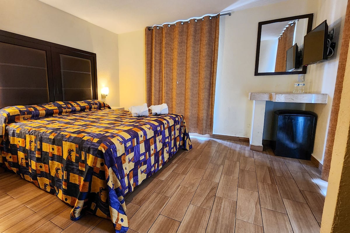 A hotel room with a large bed, a bedside table with a lamp, a small table with a phone, a mirror, and a fireplace.
