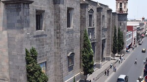 Un edificio histórico con una gran cúpula y un campanario, una calle con coches y peatones, y un balcón con barandillas ornamentales.