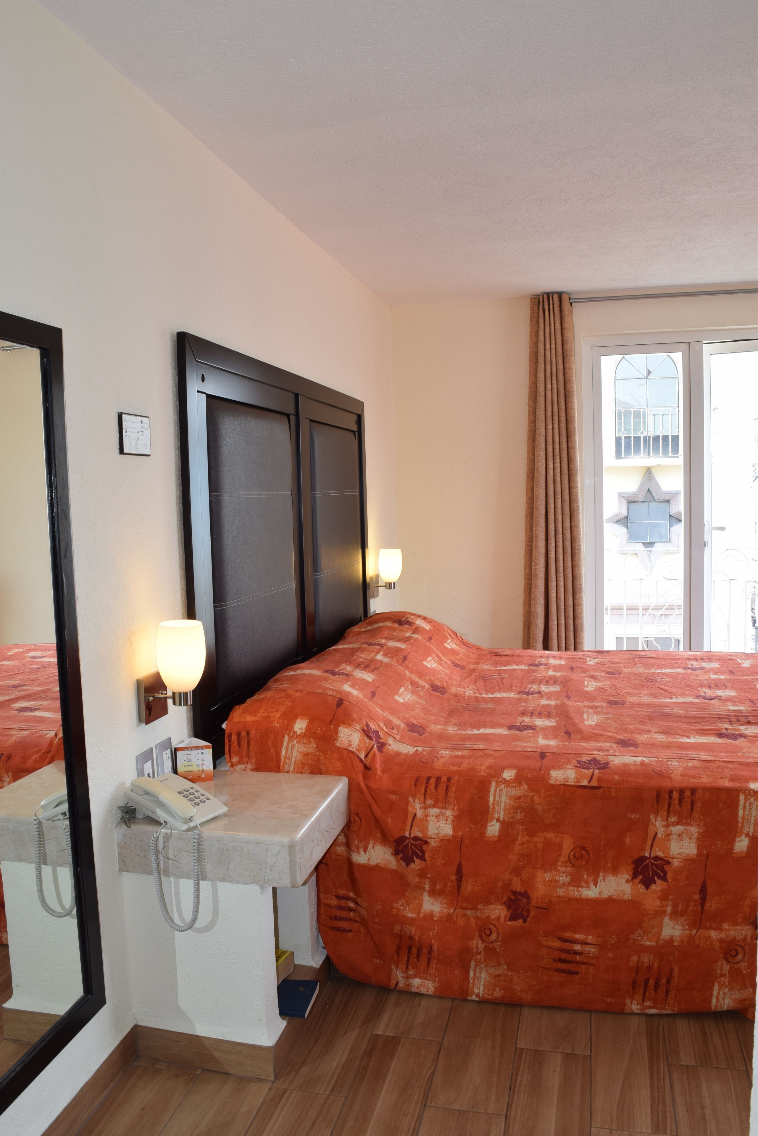 Una habitación de hotel con una cama grande, una mesita de noche con teléfono, un espejo y una ventana con cortinas.
