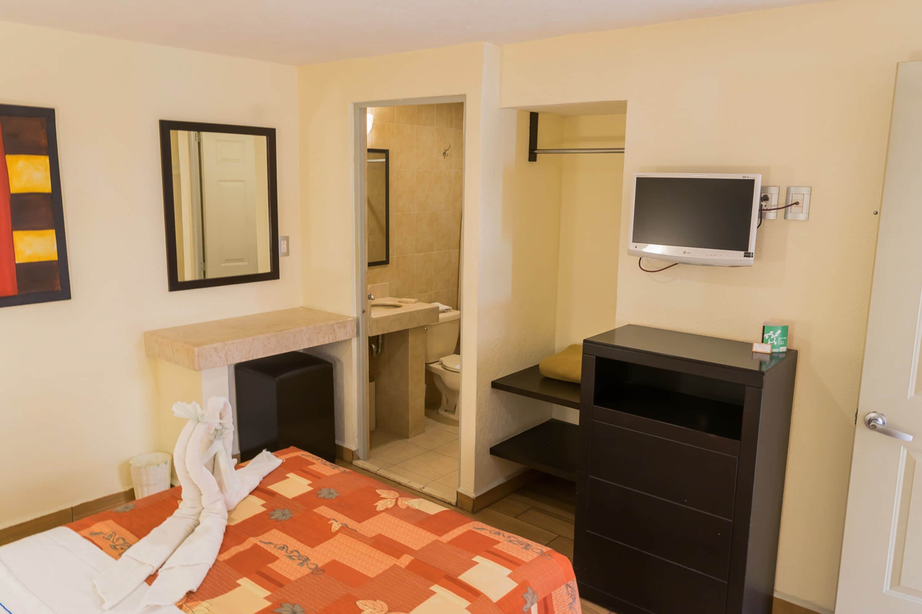 Quarto de hotel com cama, televisão, banheiro e pequena escrivaninha.